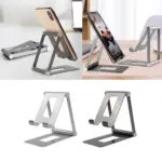 Soporte metálico plegable para celular y tablet, color plateado y gris oscuro, con un teléfono en uso.