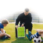 Tablero táctico de fútbol en carpeta, un entrenador explica la jugada a un niño en un campo de entrenamiento.