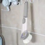 Cepillo con dispensador de jabón colgando en soporte contra pared de baldosas.