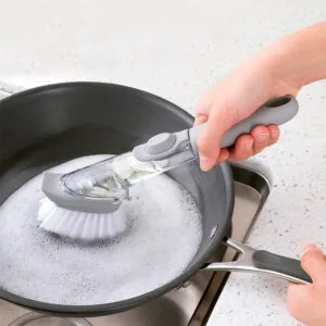 Cepillo con dispensador de jabón lavando una sartén con espuma.
