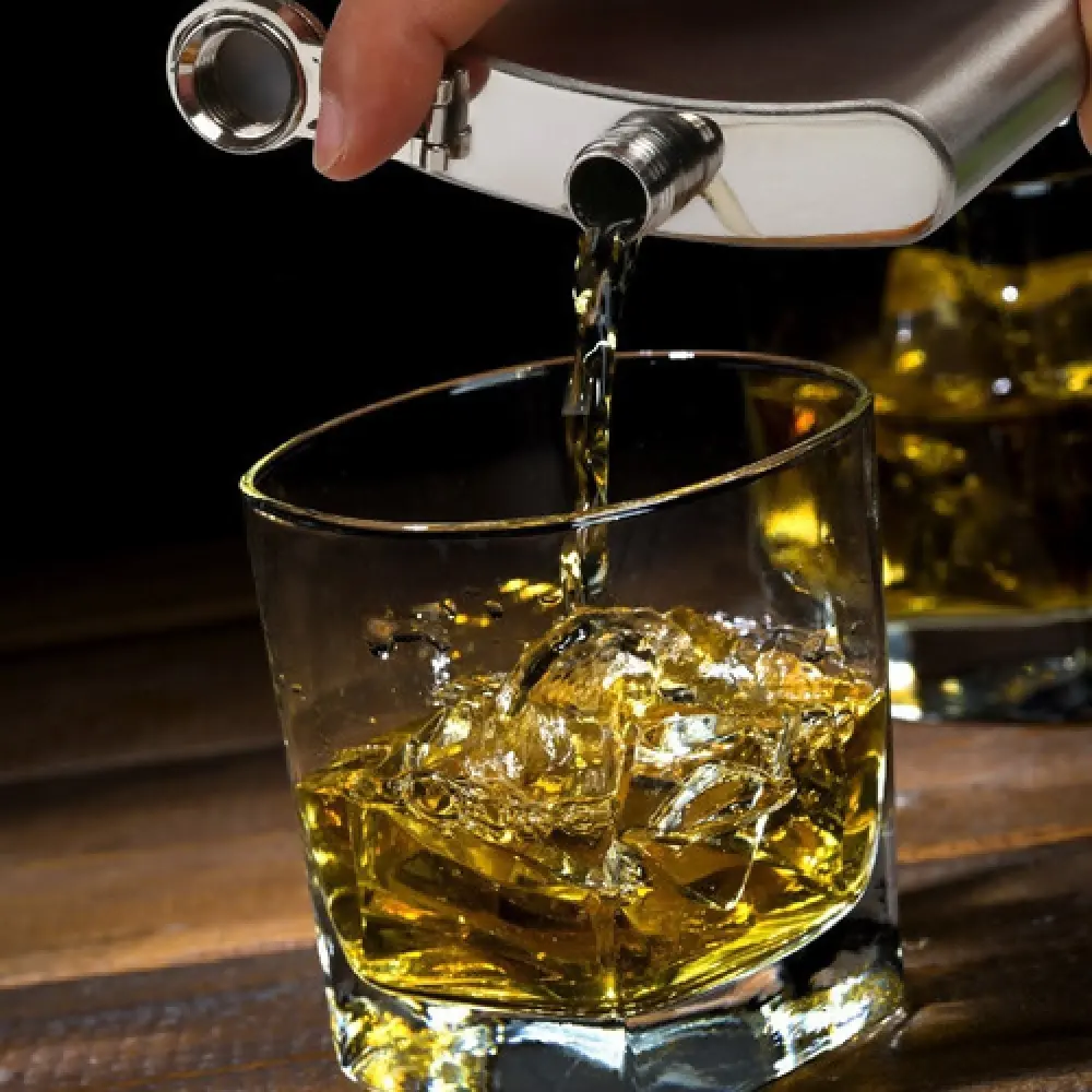 Licorera de acero inoxidable vertiendo whisky dorado con hielo en vaso.