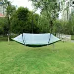 Hamaca de nailon con mosquitero, verde y blanca, suspendida entre árboles en un parque con césped.