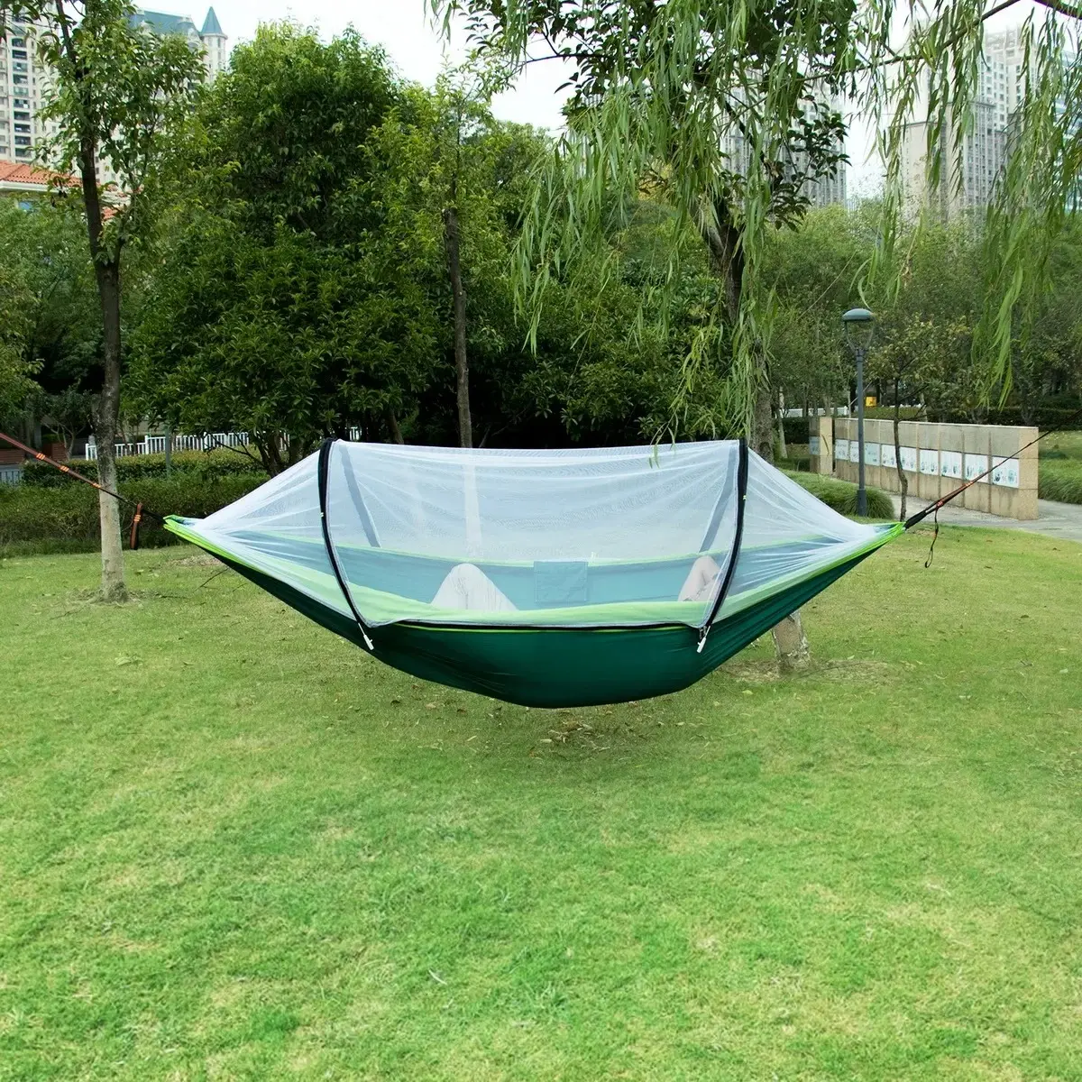 Hamaca de nailon verde con mosquitero, suspendidada entre árboles en un parque soleado.
