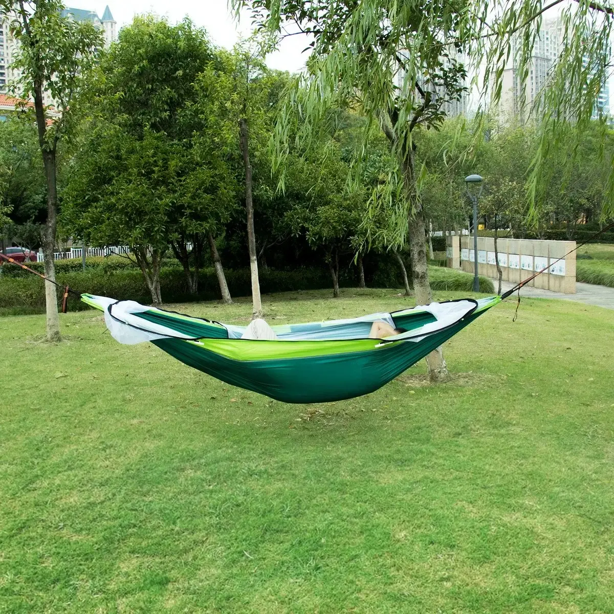 Hamaca de nailon verde y blanca con mosquitero, suspendida entre árboles en un parque.