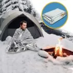 Set x2 Manta Térmica para Emergencias: Hombre envuelto en manta plateada cerca de tienda y fogata en la nieve.