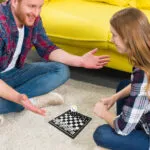 Hombre y mujer jugando mini ajedrez magnético portátil en el suelo junto a un sofá amarillo.