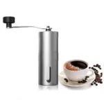 Molino manual de café en acero, molinillo portátil con manivela y taza de café al lado.