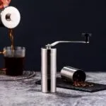 Molino de café manual de acero inoxidable con manivela y recipiente para granos.