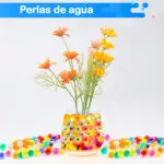 Perlas de hidrogel coloridas para decoración, en vaso con flores y esparcidas sobre superficie blanca.