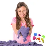 Niña sonriente jugando con arena cinética morada y moldes de playa.