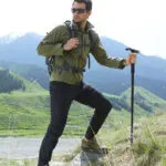Hombre con bastón de senderismo, linterna LED, chaqueta verde y mochila en montaña.