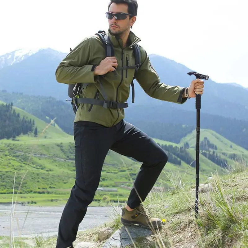 Hombre con bastón de senderismo, linterna LED, chaqueta verde y mochila en montaña.
