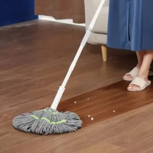 Trapeador autoescurridor gris limpiando suelo de madera, con persona en zapatillas beige al fondo.