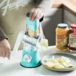 Rallador molino giratorio de verduras con base de succión rallando papas en rodajas finas sobre un plato.