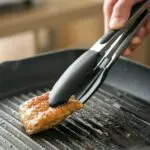 Pinzas de acero inoxidable para cocina, sujetando un trozo de carne asada en sartén grill.