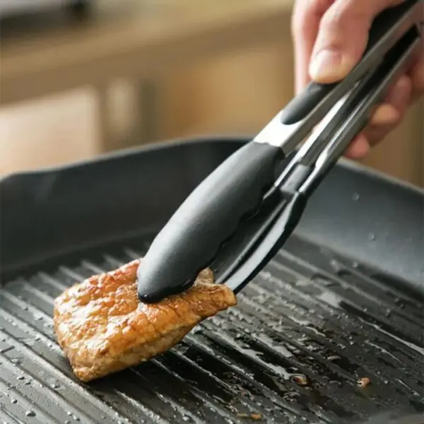 Pinzas de acero inoxidable para cocina, sujetando un trozo de carne asada en sartén grill.