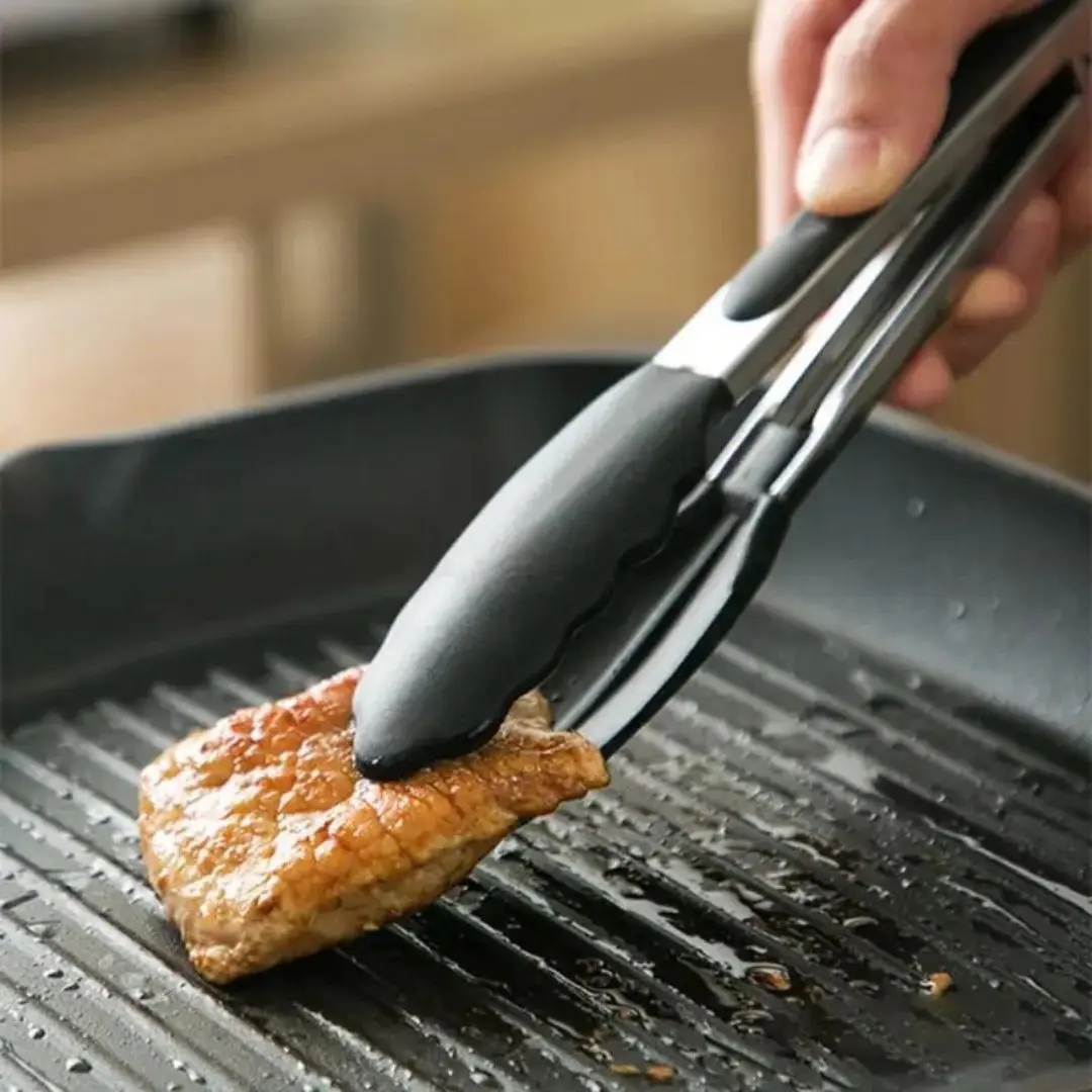 Pinzas de acero inoxidable para cocina, sujetando un trozo de carne asada en sartén grill.