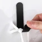 Perchero adhesivo de acero inoxidable negro con cuerda blanca, sostenido por un dedo.