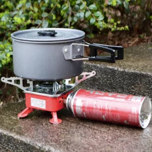 Estufa portátil a gas butano para exteriores con olla gris y patas plegables sobre piedra mojada.