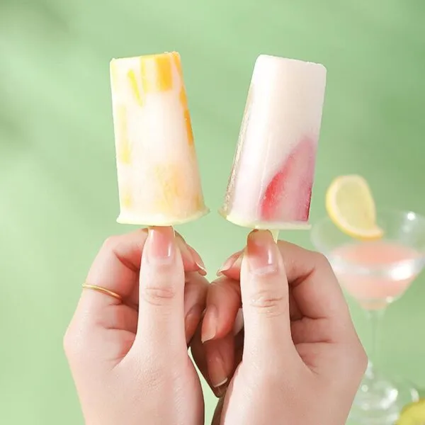 Set x2 moldes para paletas de helado con trozos de fruta, sostenidos por manos, con copa de cóctel de fondo.