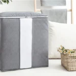 Bolsa plegable para closet gris con asas, sobre un sofá junto a canasta de flores.