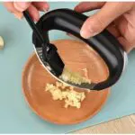 Picador ergonómico de acero inoxidable para ajo machacado en plato de madera.