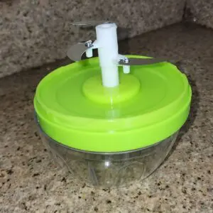 Picador de alimentos manual verde con cuchillas de acero inoxidable sobre encimera de granito.