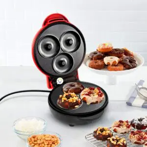 Máquina de mini donas rojas cocinando donuts glaseados y con toppings de maní y frutos rojos.