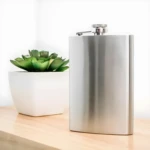 Licorera de acero inoxidable de 5 oz, diseño liso con tapa rosca, junto a planta suculenta.