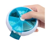 Pastillero semanal giratorio blanco y azul con pastillas verdes y blancas, sostenido por mano.