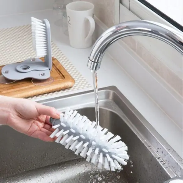Cepillo limpiador de vasos para botellas y copas, con cerdas blancas y soporte gris junto al lavaplatos.