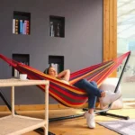 Mujer relajada en hamaca de líneas múltiples colores con soporte, junto a mesa auxiliar y estantes.