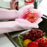 Guantes mágicos MG rosados limpiando una manzana con burbujas en un fregadero con frutas.