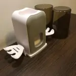 Soporte doble para cepillos y dispensador de crema dental blanco con forma de ala, junto a vasos oscuros.