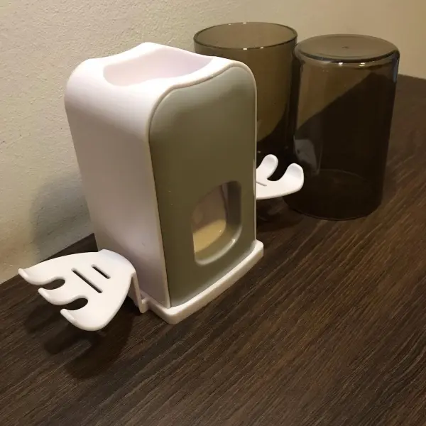 Soporte doble para cepillos y dispensador de crema dental blanco con forma de ala, junto a vasos oscuros.