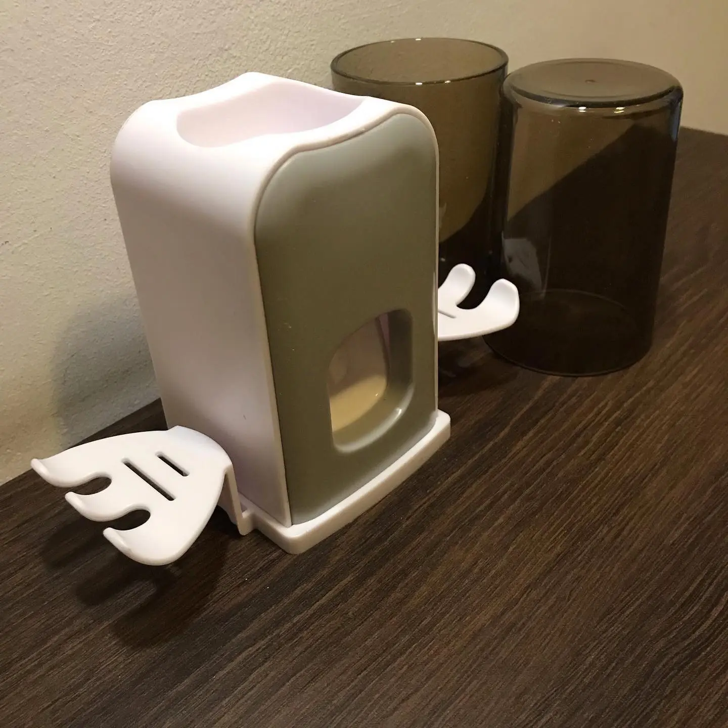 Soporte doble para cepillos y dispensador de crema dental blanco con forma de ala, junto a vasos oscuros.