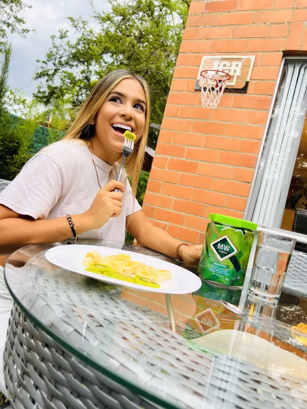 Mujer sonriente come kiwi y banana en mesa de jardín, al lado de producto Fibra Mw.