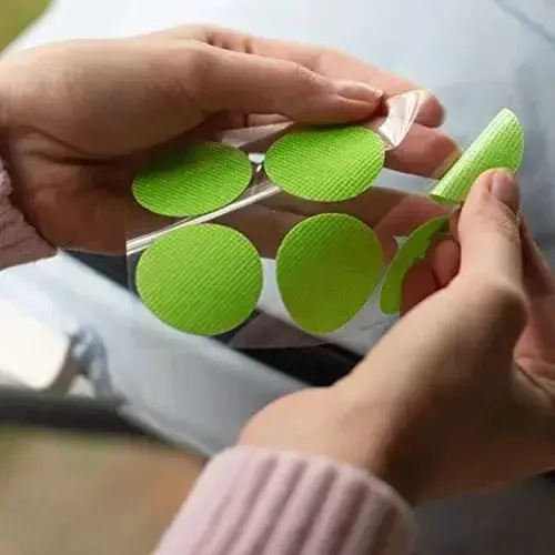 Manos aplicando parches antimosquitos de citronela redondos y verdes sobre una superficie transparente.