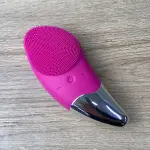 Masajeador facial limpiador rosa con cerdas de silicona y detalles metálicos plateados sobre fondo de madera.