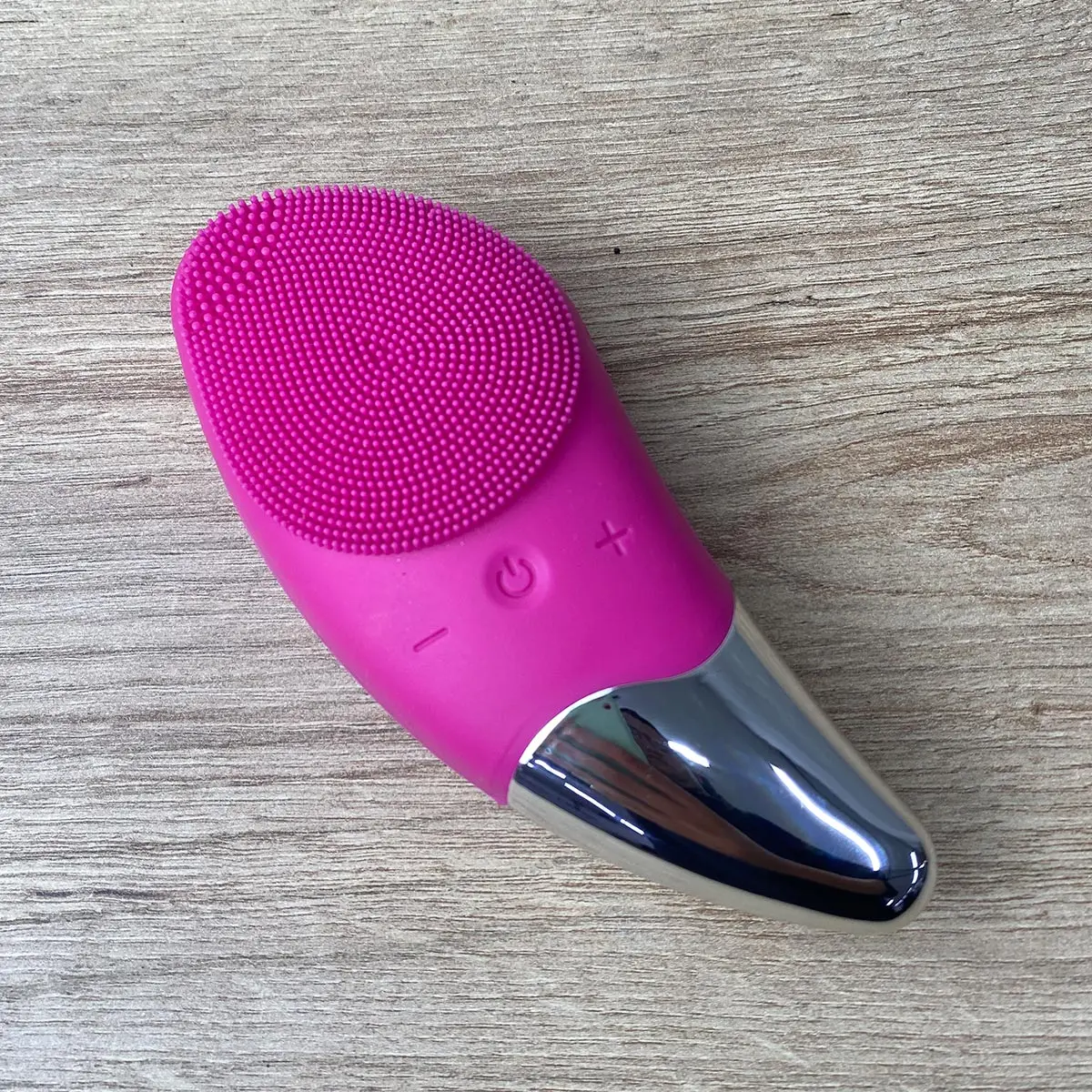 Masajeador facial limpiador rosa con cerdas de silicona y detalles metálicos plateados sobre fondo de madera.