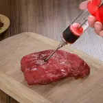 Inyector de carne con jeringa para marinar cortes de res en tabla de madera.