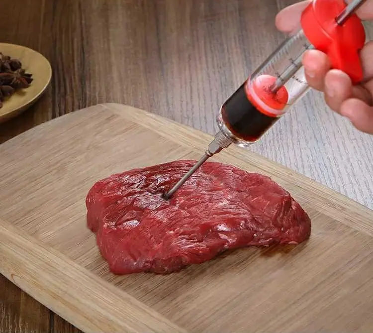 Inyector de carne con jeringa para marinar cortes de res en tabla de madera.