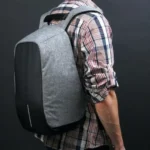 Morral antirrobo gris y negro para hombre, con fondo oscuro y camisa a cuadros.
