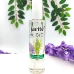 Botella de Fijador de Maquillaje Karité Bio Lemongrass con hojas verdes y flores moradas.