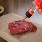 Inyector de carne inyectando líquido en un trozo de carne cruda sobre tabla de madera.