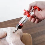 Inyector de carne con jeringa para marinar pollo crudo en mesa de madera.