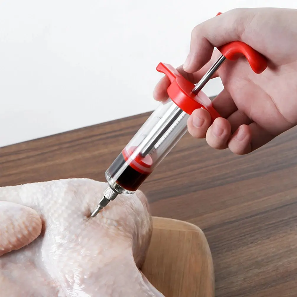 Inyector de carne con jeringa para marinar pollo crudo en mesa de madera.