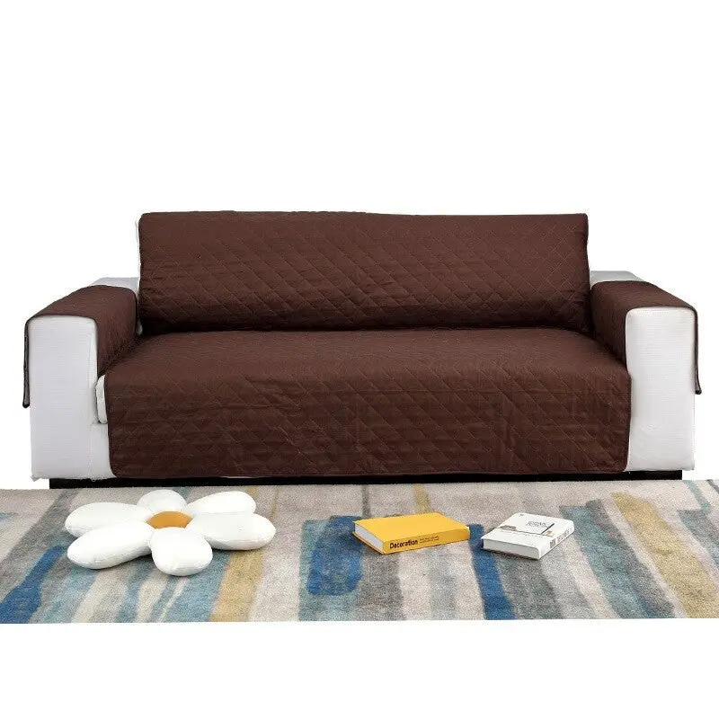 Cubre sofá marrón de 3 puestos, acolchado con rombos, sobre sofá blanco, con cojín en forma de flor y libros en alfombra.