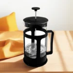 Cafetera prensa francesa con filtro de acero y jarra de vidrio, sobre mesa de madera con cojín naranja.