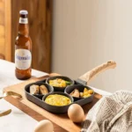 Sartén antiadherente para 4 huevos con comida variada y cerveza Hoegaarden en tabla de madera.